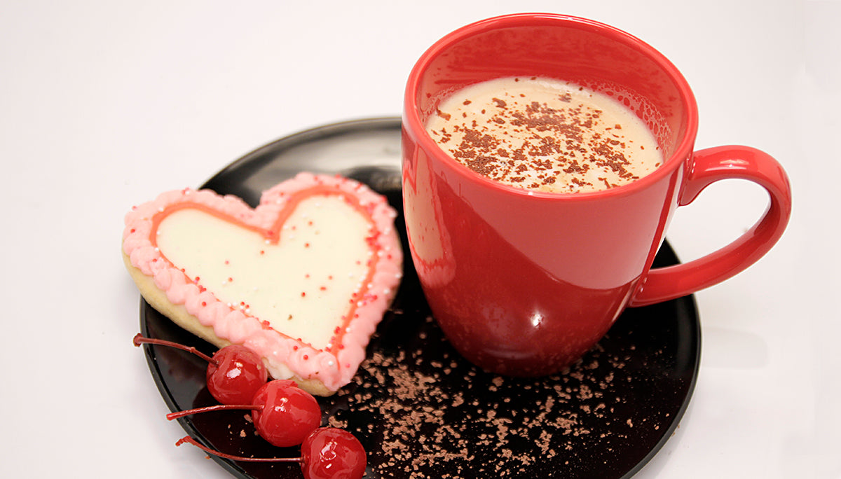 A red mug and heart shaped sugar coffee on a plate. Coffee with cinnamon like topping  on top of coffee. Three red cherries are garnishing the plate.