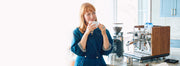 Woman holding a coffee cup in a kitchen with an ECM Synchronika Espresso Machine with wood accents next to a Ceado Grinder on a marble counter. 