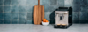 DeLonghi Magnifica EVO Super-Automatic machine on a gray counter top and in front of a blue tiled wall.