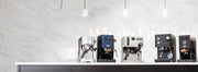 Line up of 6 best of espresso machines of 2026 on a white and black counter in front of a white wall with lights hanging above.