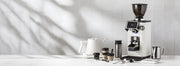 Ceado REV Zero grinder in white next to a varia smart kettle, notNeutral pico cups, a revolution milk frothing pitcher, spoons, scale and tampers on a white counter top in front of a white tiled wall.