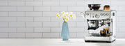 Breville Barista Express Impress in Stainless Steel on a white counter top next to flowers and in front of a white tiled wall.