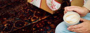 Person sitting on a patterned rug holding a latte cup with latte art in it and gifts in the background.