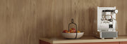 Close-up of a Stone Lite Espresso Machine in stainless steel on a wooden counter top with salmon colored cupboards next to a bowl of apples in front of a wooden wall.