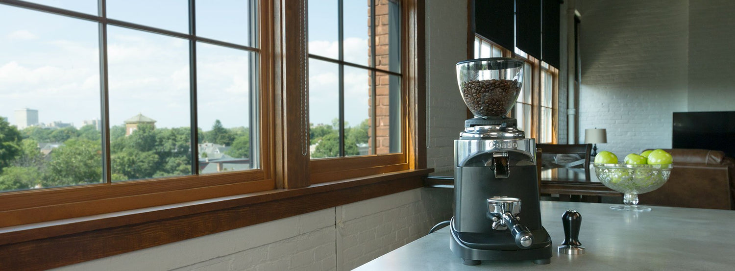 A Ceado coffee and espresso grinder at the edge of a white countertop, next to a large window.