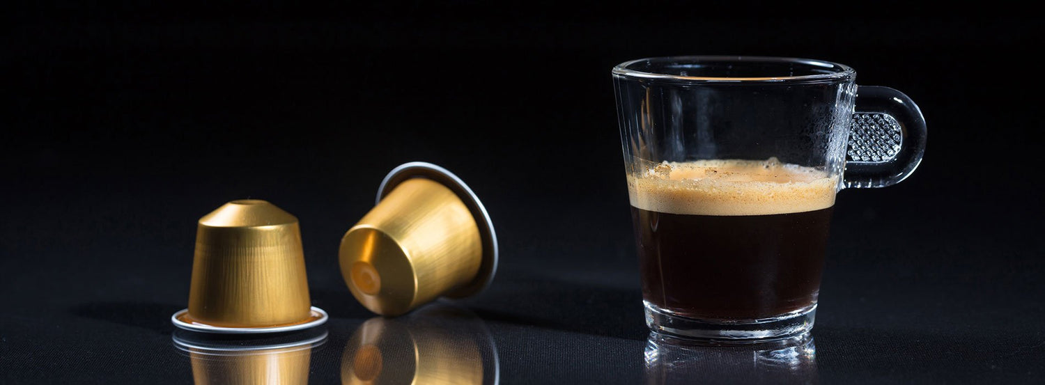 A cup of coffee made with a single serve coffee maker, next to two golden coffee pods.