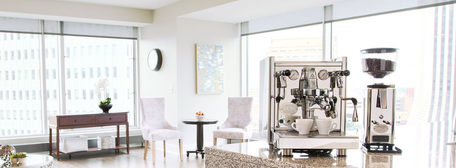 A mostly white room with floor to ceiling windows, and an espresso machine and coffee grinder package sitting on a granite countertop.