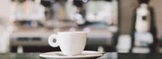 A white cup and saucer in front of an out of focus espresso machine and coffee grinder.
