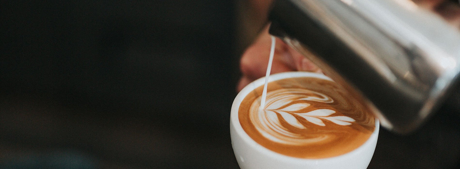 Milk pouring into a coffee cup to make latte art of a frond on the surface.