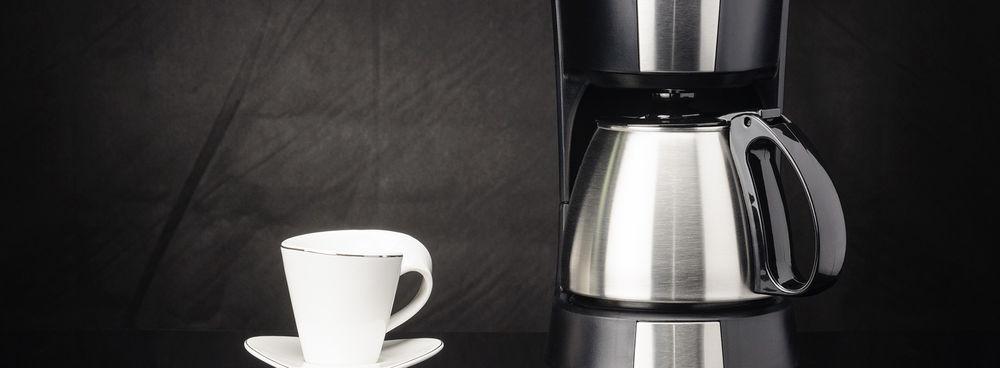 A stainless steel coffee maker and thermal carafe next to a white cup and saucer