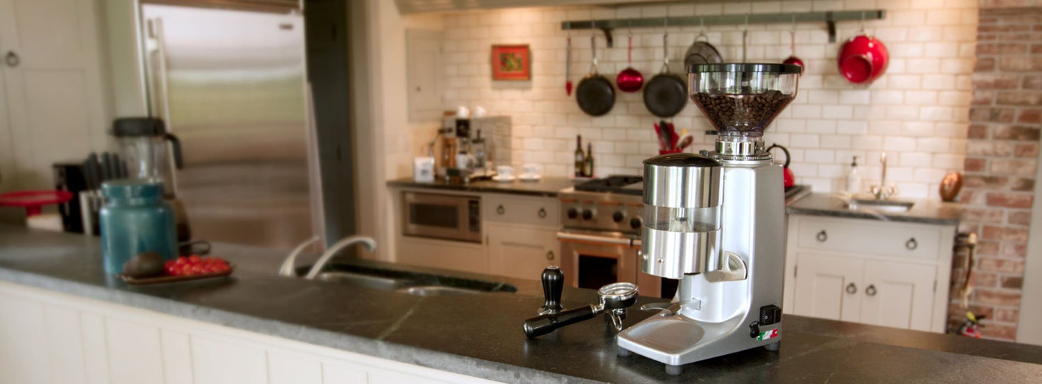 A Quamar coffee and espresso grinder on a granite counter with pots and pans hanging up on the white brick wall in the background.