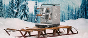 Close-up of the Profitec JUMP Espresso Machine on a wooden sled in front of a winter forest scene with snowfall.