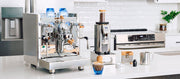 Close-up of a coffee setup bundle in a bright kitchen setting, featuring a stainless steel espresso machine, a coffee grinder, blue cups, and a prepared coffee cup on a white countertop.