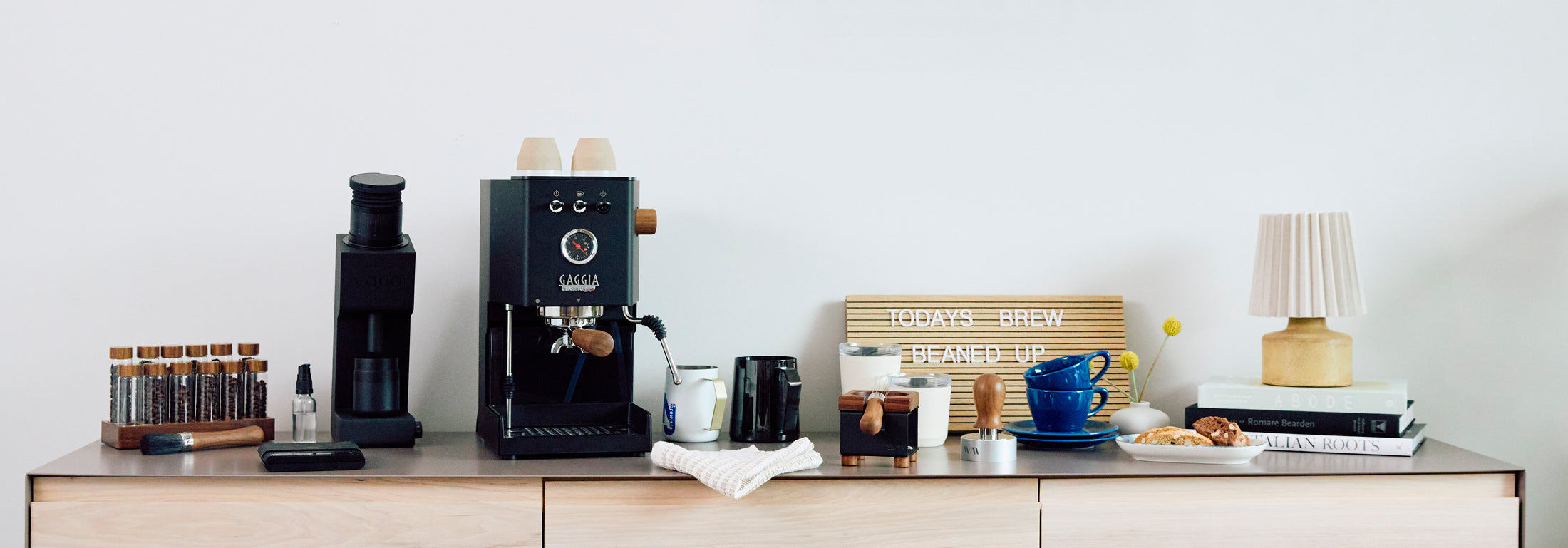 Gaggia Classic UP on a wooden surface next to the Varia VS4 in Black and other different coffee accessories and cups in front of a white background.