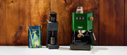 Close-up of the Gaggia Classic E24 in Green next to a Varia Grinder in black and Whole Latte Love beaned up coffee in front of a wooden wall.