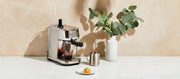 Close-up of a Breville Bambino espresso machine on a table with a vase of eucalyptus and a plate of food.