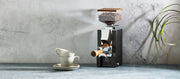 Close-up of a Eureka Mignon Silenzio 55 in Black with Walnut accents on a gray stone counter and gray background next to latte cups.