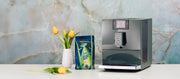 Close-up of the Jura C9 espresso machine next to a package of Whole Latte Love Beaned UP and flowers on a white counter in front of a marble background.