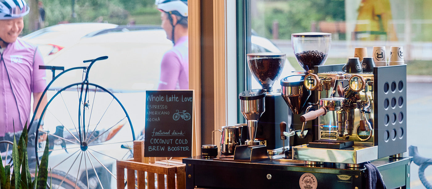 Close-up of the Rocket Espresso Appartamento TCA in black next to a Rocket Espresso Faustino and FAUSTO grinder with cyclists in the background.