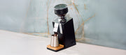 Close-up of the Eureka Speedy 65 in Black grinding beans on a white counter in front of a blue slate wall.