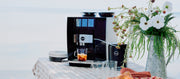 Close-up of a JURA Super Automatic Espresso machine on a picnic table next to cups plates and flowers.