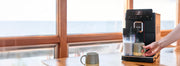 Gaggia Magenta Prestige brewing a latte into a green mug on a table in front of a window.