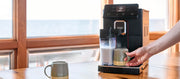 Close-up of the Gaggia Magenta Prestige brewing a latte into a green mug on a table in front of a window.