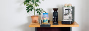 Stone Lite espresso machine next to a Baratza grinder and Whole Latte Love Buzzopolis coffee on a wooden counter top.
