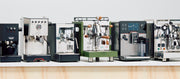 Close-up of a row of various espresso machines on a white surface with a white background.