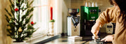 Holiday themed kitchen with a Green Gaggia Classic Espresso Machine next to a Gaggia MDF Grinder with a woman preparing a shot of espresso.