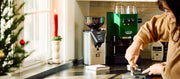 Close up of a holiday themed kitchen with a Green Gaggia Classic Espresso Machine next to a Gaggia MDF Grinder with a woman preparing a shot of espresso.