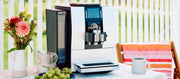 Close-up of the silver JURA Z6 espresso machine on an outdoor table, with a cup of coffee and a small bouquet of flowers beside it.