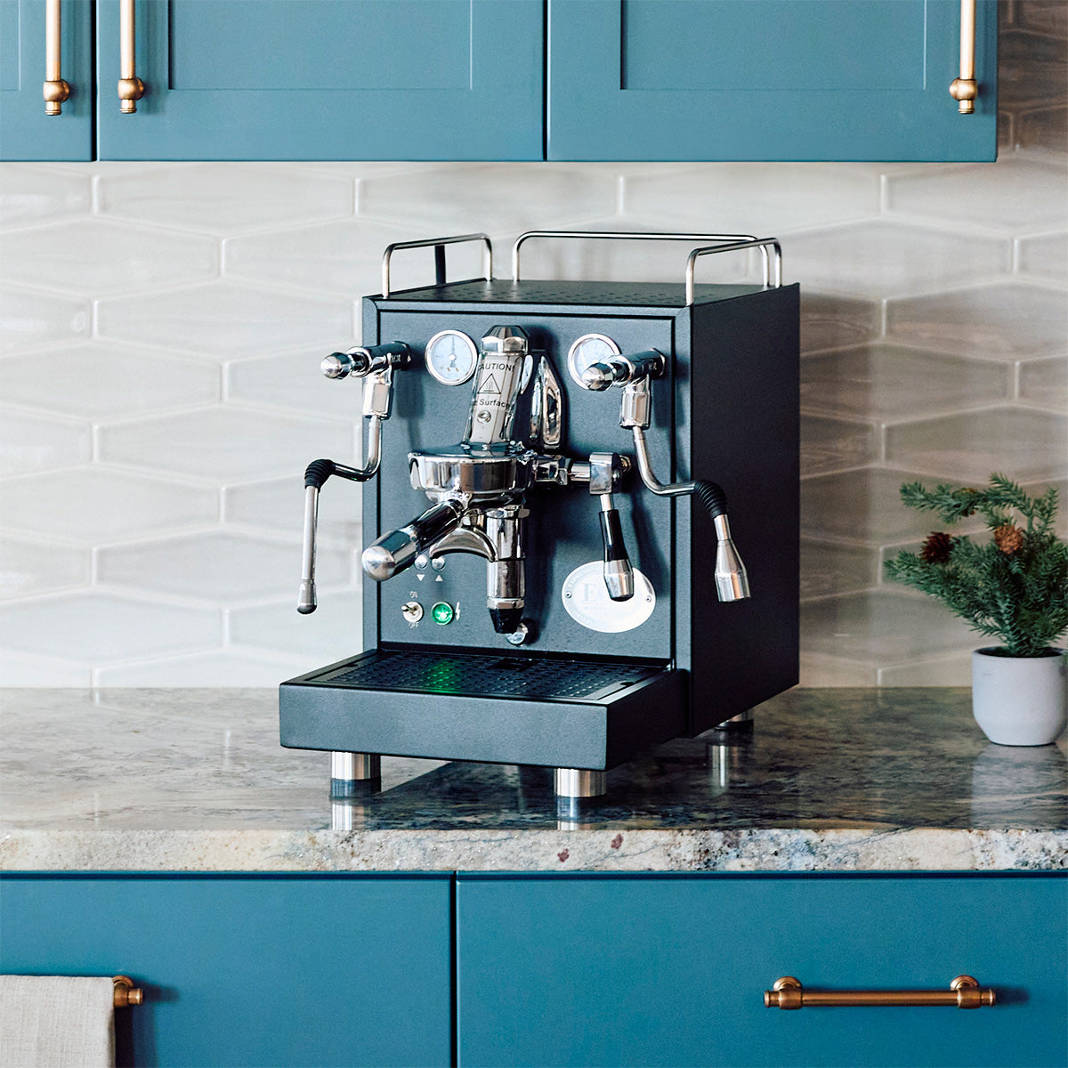 Black ECM Mechanika Max Espresso machine on a kitchen counter with blue cabinets and a plant in the background - Machine Giveaway.