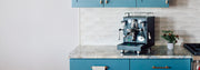 Espresso machine on a kitchen counter with blue cabinets and white tiled wall.