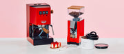 Close-up of the Cherry Red Gaggia Classic E24 next to a Red Eureka Silenzio 55 in front of a pink background and on a white counter.