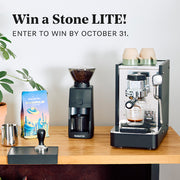Stone Lite espresso machine next to a Baratza grinder and Whole Latte Love Buzzopolis coffee on a wooden counter top. Promotional text that reads "Win a Stone Lite! Enter to win by October 31."