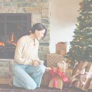 Woman opening a gift in front of a fireplace and Christmas tree.