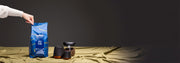 Person holding bag of Lavazza Super Crema Coffee next to coffee cups and canisters in front of a black wall.