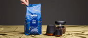 Close-up of Person holding bag of Lavazza Super Crema Coffee next to coffee cups and canisters in front of a black wall.