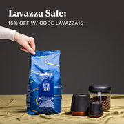 Person holding bag of Lavazza Super Crema Coffee next to coffee cups and canisters in front of a black wall with promotional text that reads "Lavazza Sale".