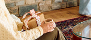 Person sitting on a couch holding a mug and wrapped gift in a cozy living room.