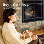 Woman sitting by a fireplace holding a wrapped gift and a mug, with text overlay for a sale promotion that says "Buy 3 Get 1 Free".
