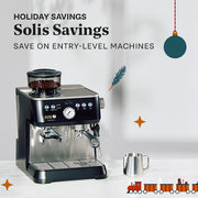 Black Solis espresso machine sitting on a white counter next to a frothing pitcher with holiday decorations.