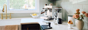 Bezzera NINI in white with wooden accents next to a Eureka Compatto Grinder in white with wood accents and other coffee accessories, flowers and a sink in front of a window on a white counter top with a sage backsplash. 