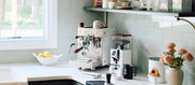 Close-up of a Bezzera NINI in white with wooden accents next to a Eureka Compatto Grinder in white with wood accents and other coffee accessories, flowers and a sink in front of a window on a white counter top with a sage backsplash. 