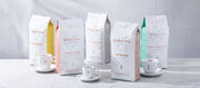 Close-up of Caffe Carraro coffee bags lined up with Carraro espresso cups on a white counter top in front of a white tiled wall.