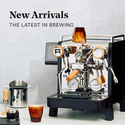 Close-up of the Rocket Espresso R58 Tune in Black with Olive Wood Accents next to coffee accessories on a white counter in front of a white wall. Promotional text overlayed that reads "New Arrivals - The Latest in Brewing".