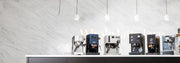 Line up of 6 best of espresso machines of 2026 on a white and black counter in front of a white wall with lights hanging above.