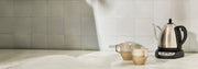A stainless steel electric kettle with a black handle and base sits on a countertop next to two beige ceramic mugs against a light gray tiled wall.