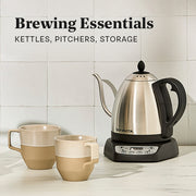 A stainless steel electric kettle with a black handle and base sits on a countertop against a white tiled wall. Two ceramic mugs in beige and cream are placed in front. Text overlayed that reads "Brewing Essentials - Kettles, Pitchers, Storage".